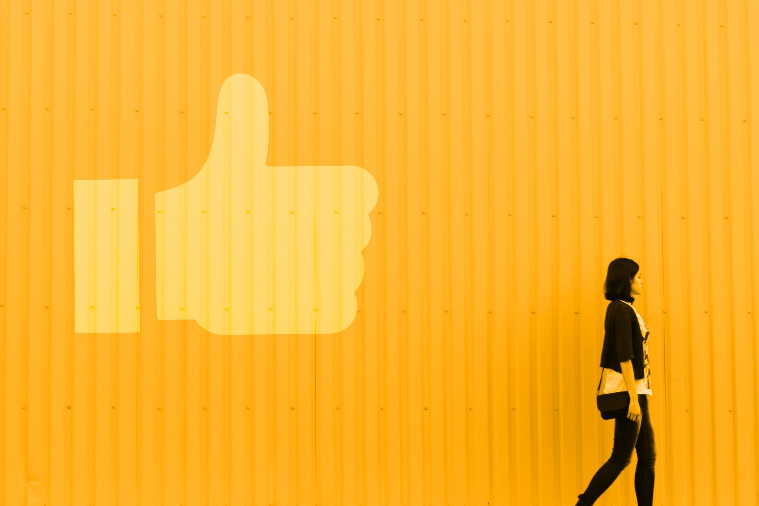 woman walking past large facebook like icon yellow wall