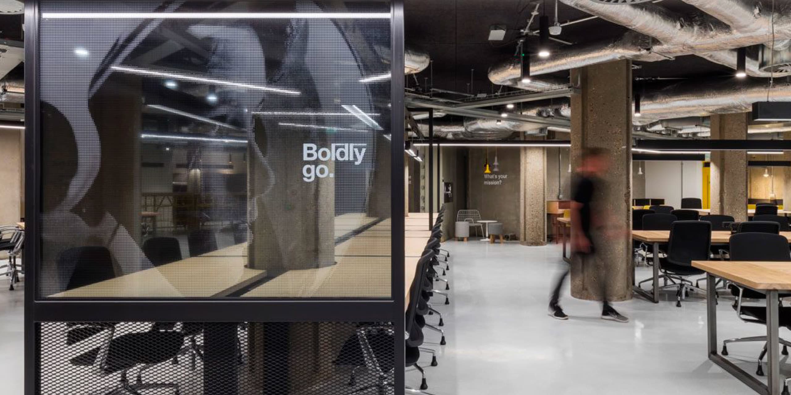 Open-plan workspace with “Boldly go.” graphic on a glass partition and long bench tables
