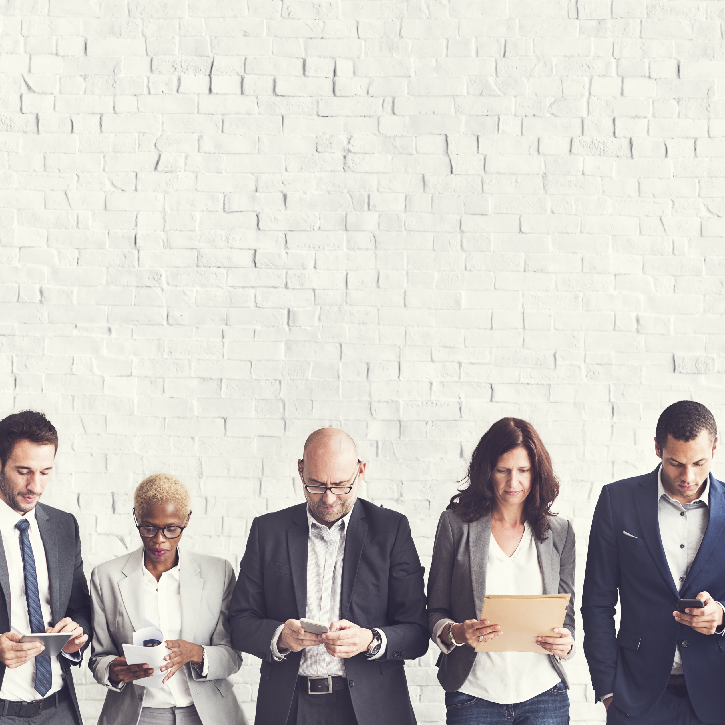 Five business professionals standing against a white brick wall, looking at phones and documents