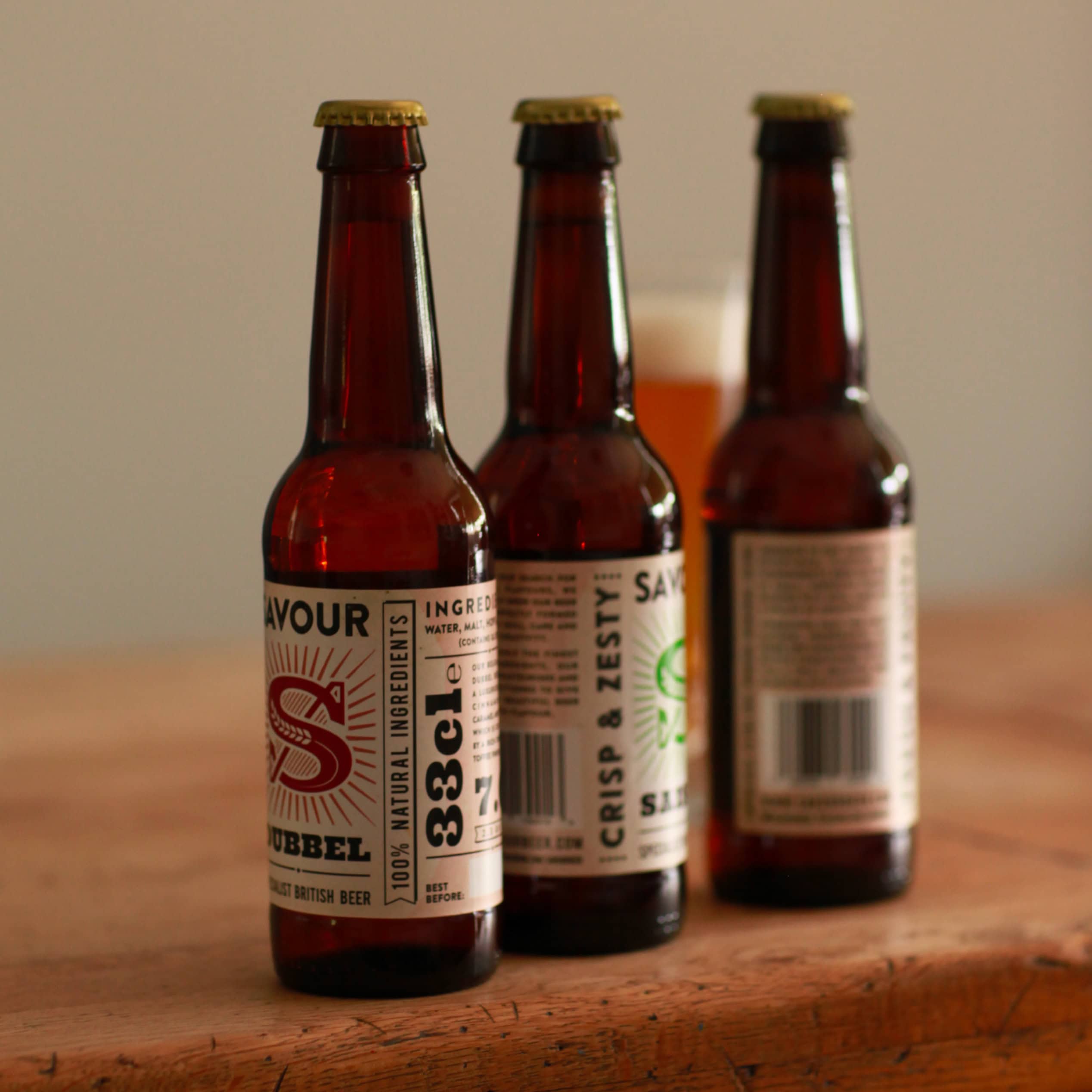 Three Savour beer bottles on a wooden table with a glass of beer in the background