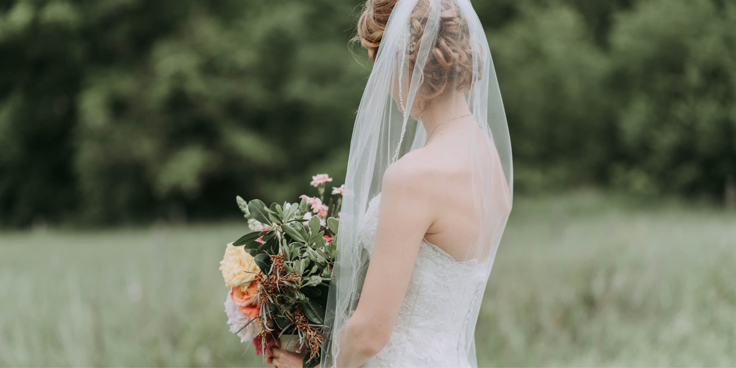 Bride in a lace dress and veil holding a bouquet in a meadow