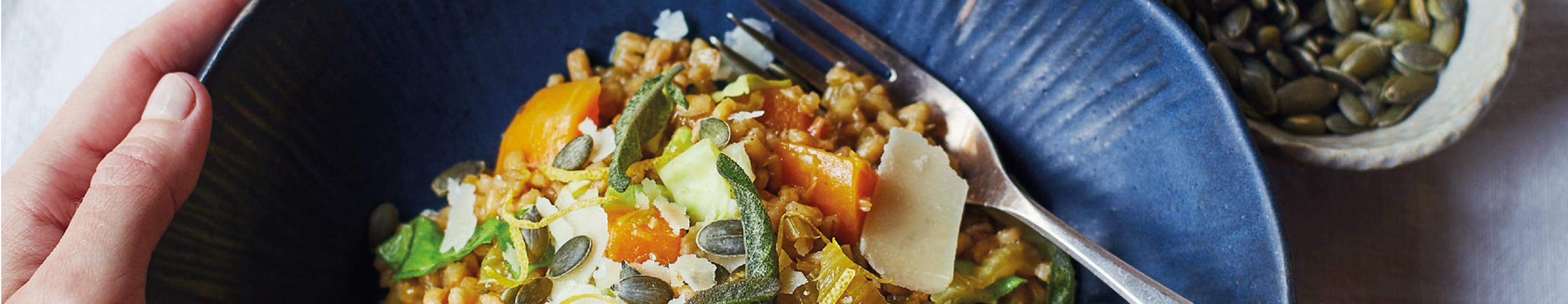 Overhead crop of a grain bowl topped with herbs, seeds and shaved cheese, with hands holding the bowl and fork