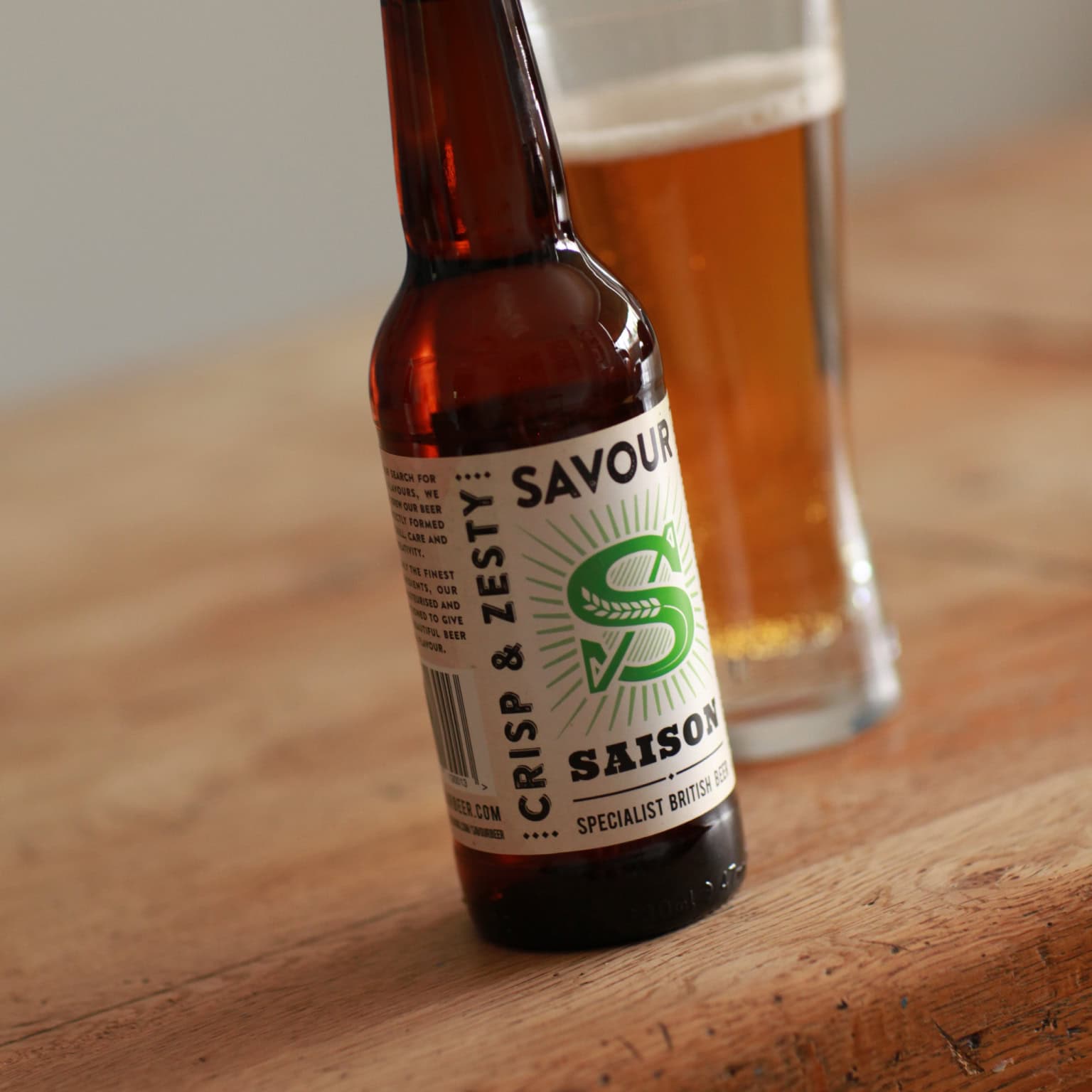 Bottle of Crips & Zesty Savour Saison beer on a wooden table with a pint glass in the background.