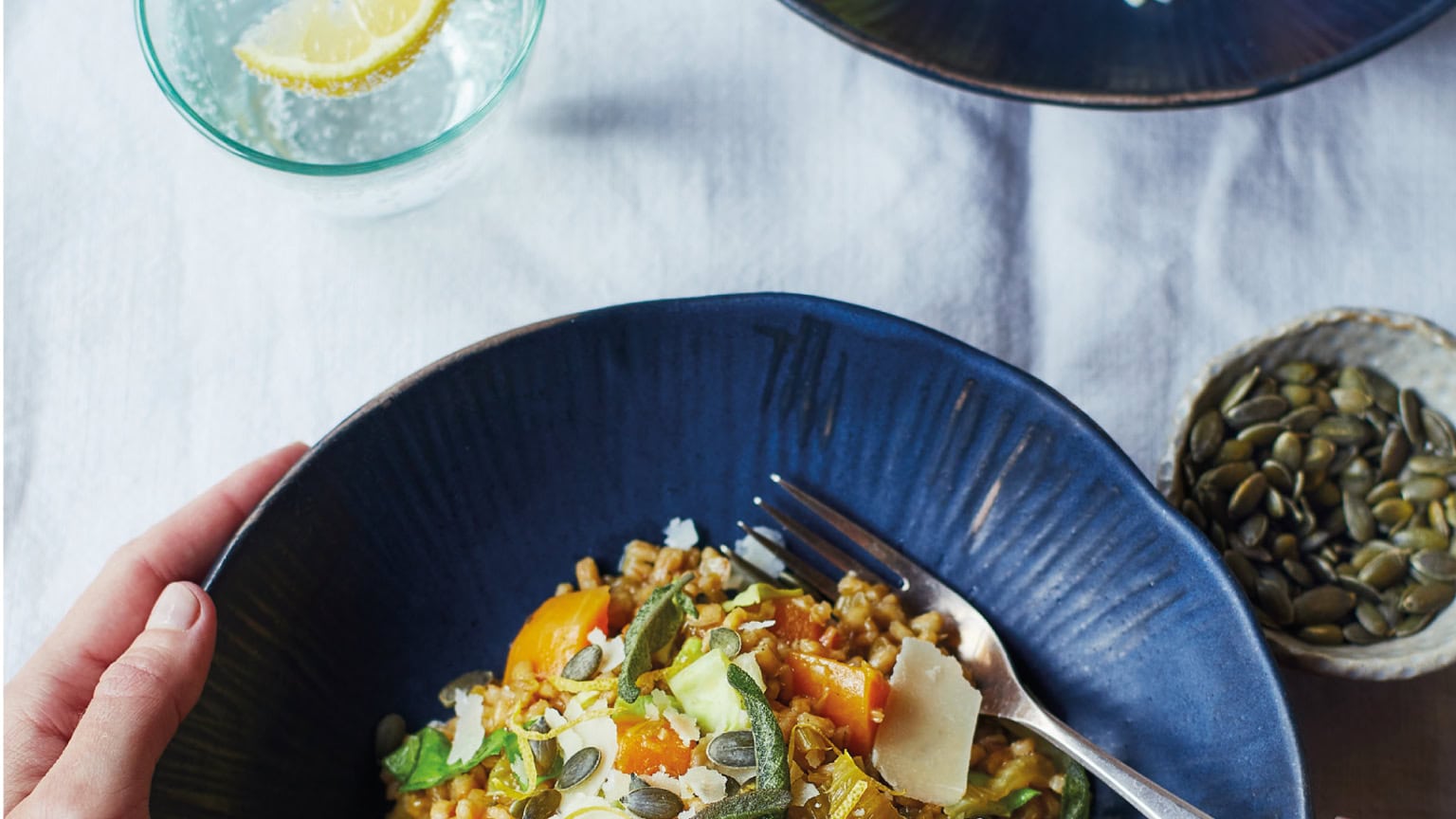 Hand holding a blue bowl of grain salad with squash, shaved cheese and pumpkin seeds on a white tablecloth.